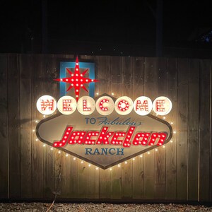K&ouml;nnte beinhalten: Ein beleuchtetes Schild im Vintage-Stil mit wei&szlig;en Lichtern und roten Gl&uuml;hbirnen, die "WELCOME TO Fabulous Jackelam RANCH" buchstabieren. Das Schild hat die Form eines Diamanten mit einem Stern oben drauf.