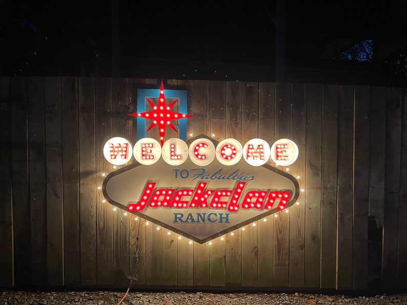 K&ouml;nnte beinhalten: Ein beleuchtetes Schild im Vintage-Stil mit wei&szlig;en Lichtern und roten Gl&uuml;hbirnen, die "WELCOME TO Fabulous Jackelam RANCH" buchstabieren. Das Schild hat die Form eines Diamanten mit einem Stern oben drauf.