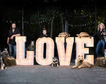 Könnte beinhalten: Eine Familie posiert vor einem großen beleuchteten Schild, das "LOVE" buchstabiert. Die Familie besteht aus zwei Erwachsenen, einem jungen Mädchen und mehreren Hunden. Das Schild ist mit Glühbirnen verziert. Die Familie steht in einem Waldgebiet mit Lichterketten, die über ihnen hängen.