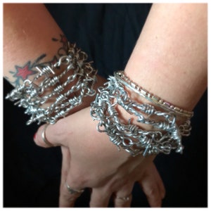 May include: A set of silver barbed wire bracelets. The bracelets are stacked on each other and are worn on both wrists.