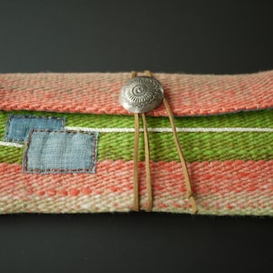 Op de afbeelding: Een geweven clutch met een groen, roze en wit gestreept patroon. De clutch heeft een klep sluiting en een zilveren concho met een bloemenmotief. De clutch heeft een bruin leren band.