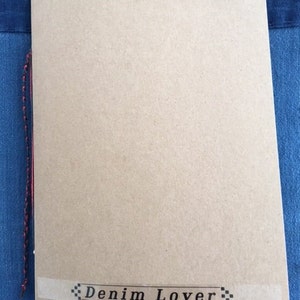Peut inclure: Un carnet marron avec un cordon rouge attaché à la reliure. Le carnet a une étiquette blanche avec le texte "Denim Lover" en noir.