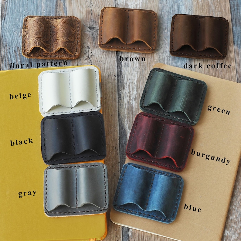 May include: A set of ten leather pen holders in various colors, including beige, black, gray, brown, dark coffee, green, burgundy, and blue. The holders are all made of leather and have a book-like design. The holders are displayed on a brown surface.