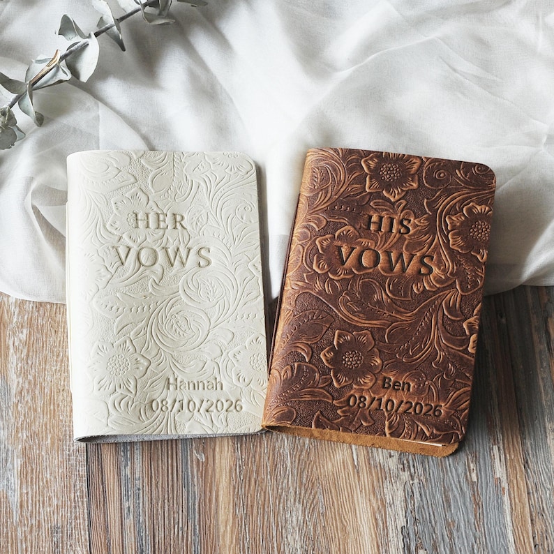 May include: Two leather-bound vow books, one cream and one brown. The cream book reads "HER VOWS" with the name "Hannah" and the date "08/10/2026". The brown book reads "HIS VOWS" with the name "Ben" and the date "08/10/2026".