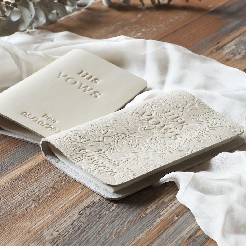 May include: Two off-white vow books with embossed text and floral patterns. One book reads "HIS VOWS" with a date. The other book reads "HER VOWS" with a date and name. The books are on a wooden surface.