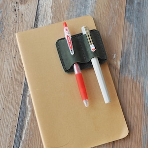 May include: A dark green leather pen holder with two pens inserted. The pen holder has two slots for pens. One pen is red and the other is white.