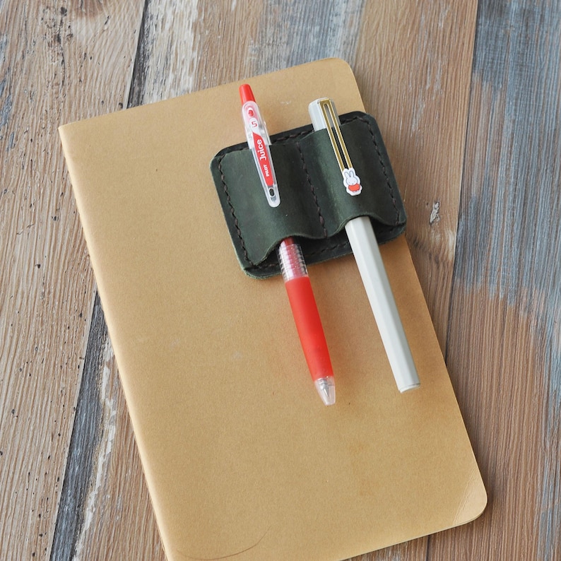 May include: A dark green leather pen holder with two pens inserted. The pen holder has two slots for pens. One pen is red and the other is white.