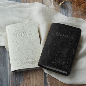 May include: Two wedding vow books, one white and one black, with floral embossed designs. The word "VOWS" is embossed on the cover of each book. The white book is personalized with the name "Emma" and the date "09/05/2023". The black book is personalized with the name "Dalton" and the date "09/05/2025".