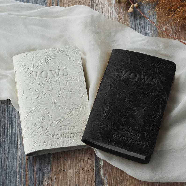 May include: Two wedding vow books, one white and one black, with floral embossed designs. The word "VOWS" is embossed on the cover of each book. The white book is personalized with the name "Emma" and the date "09/05/2023". The black book is personalized with the name "Dalton" and the date "09/05/2025".
