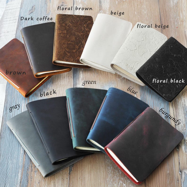 May include: A collection of leather journals in various colors, including brown, black, gray, green, and blue. Some journals feature embossed floral patterns. The journals are open, revealing the pages.