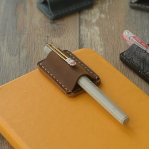 Könnte beinhalten: Ein brauner Stiftehalter aus Leder mit sichtbaren weißen Nähten, befestigt an einem leuchtend gelben Notizbuch. Ein hellgrauer Stift steckt im Halter und hat einen goldenen Clip mit einem kleinen weißen und orangen Hasensymbol oben.