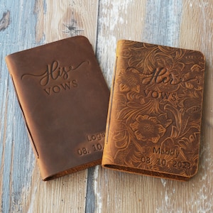 May include: Two brown leather vow books with embossed floral designs. The book on the left says "His Vows" and has the date "08.10" and the word "Logan" on the bottom. The book on the right says "Her Vows" and has the date "08.10.2023" and the name "Maddy" on the bottom.