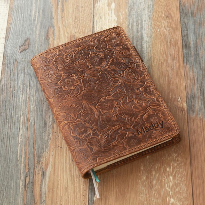 May include: A brown leather journal with an embossed floral design. The journal has the name "Maddy" engraved on the cover. The journal is closed and has a pen holder.