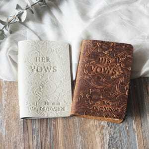 May include: Two wedding vow books, one cream-colored and one brown, with floral embossed designs. The cream book reads "HER VOWS" and the brown book reads "HIS VOWS". Both books are personalized with names and dates.