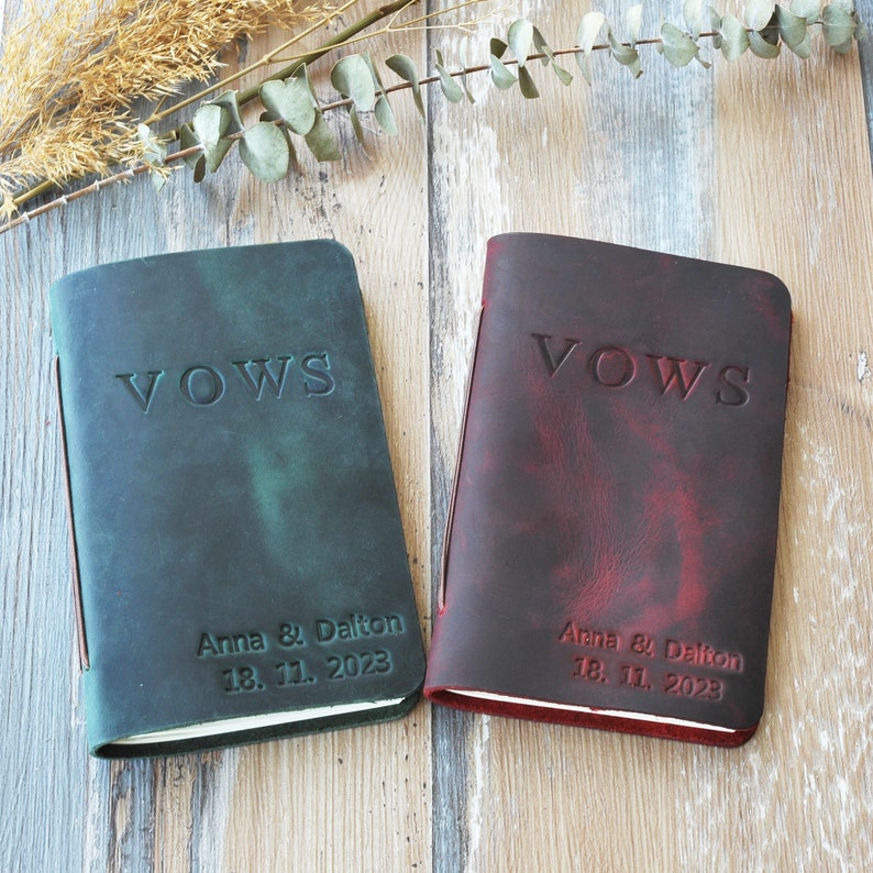 May include: Two leather-bound vow books, one teal and one burgundy, with the word "VOWS" embossed on the cover. Each book has the names "Anna & Dalton" and the date "18. 11. 2023" embossed below the word "VOWS".