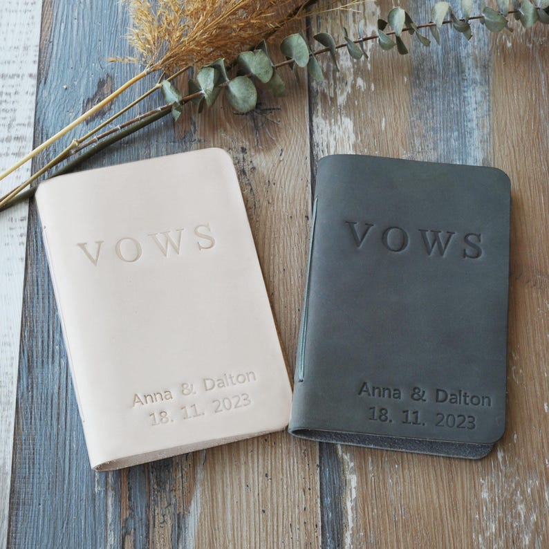 May include: Two leather vow books, one cream and one gray, lie side-by-side on a wooden surface. The word "VOWS" is embossed on each cover. The cream book has the names "Anna & Dalton" and the date "18. 11. 2023" embossed below.