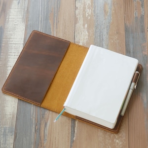 May include: Open leather journal with a brown cover and a cream-colored interior. A white notebook with a blue ribbon bookmark is inside. A pen is tucked into the journal's spine. The journal is on a wooden surface.
