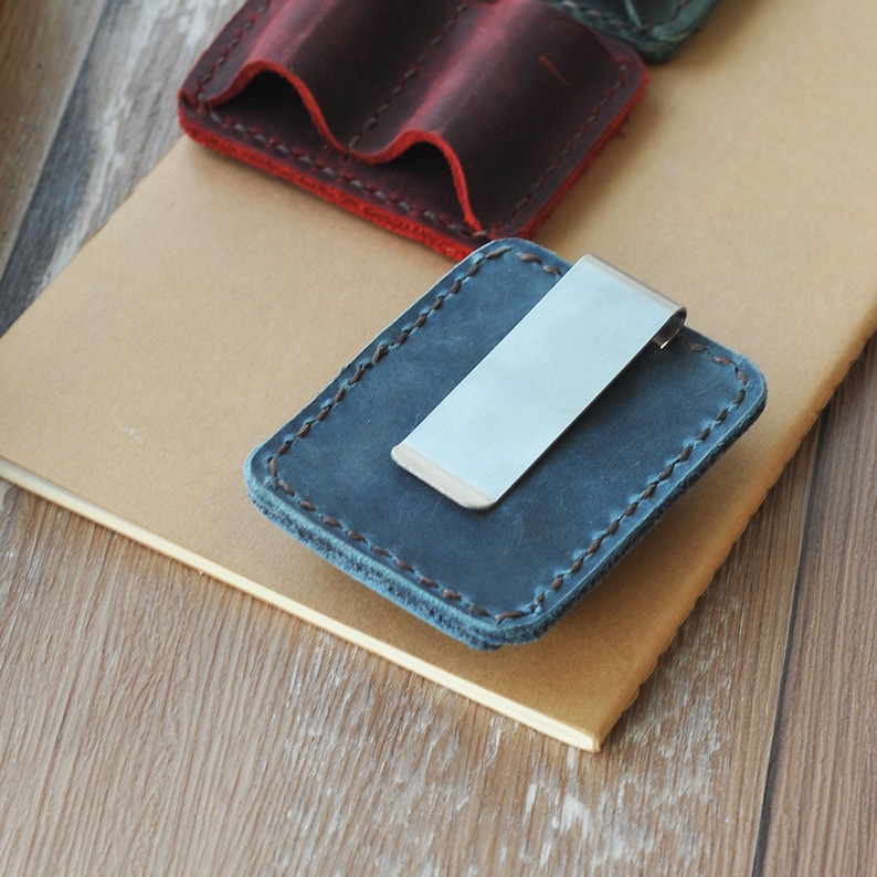 May include: A blue leather wallet with a metal money clip. The wallet has a stitched edge and is lying on a brown surface.