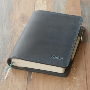 May include: A dark blue leather journal with the name "Dalton" embossed on the cover. The journal has a pen holder and a ribbon bookmark. The pages are cream-colored, and the journal is on a wooden surface.