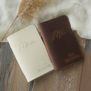 Puede incluir: Dos diarios de cuero, uno crema y otro marrón, se exhiben sobre una superficie de madera. El diario crema está grabado con "Mrs", "Rachel" y "16/10/2026". El diario marrón está grabado con "Mr", "Chris" y "16/10/2026".