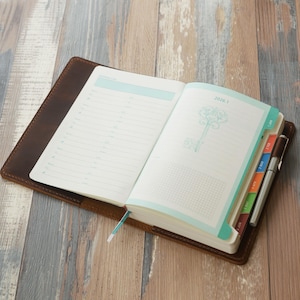 May include: Open leather-bound journal with lined pages and a key design on the right page. The journal has a light blue border and a bookmark. A pen rests on the right side of the journal.