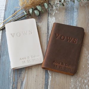 May include: Two leather vow books, one cream and one brown, with embossed text. The word "VOWS" is at the top, and the names "Anna & Dalton" with the date "18. 11. 2023" are below. The books are on a wood-grain surface.
