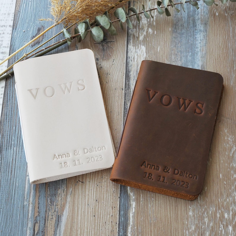 May include: Two leather vow books, one cream and one brown, with embossed text. The word "VOWS" is at the top, and the names "Anna & Dalton" with the date "18. 11. 2023" are below. The books are on a wood-grain surface.