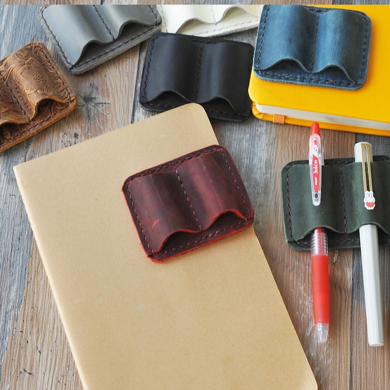 May include: A close-up of a brown leather notebook with a red leather pen holder attached to the cover. The notebook is lying on a wooden surface. There are other leather pen holders in the background.