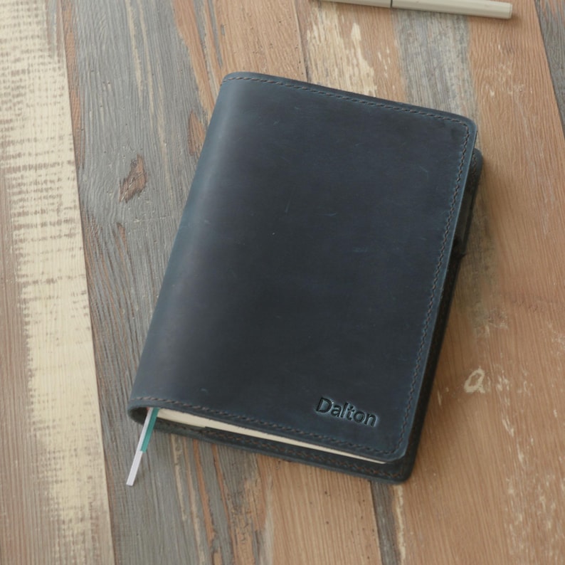 May include: A dark blue leather journal with the name "Dalton" embossed on the cover. The journal has a sewn edge and a light green ribbon bookmark. The journal is lying on a wooden surface.