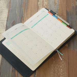 May include: Open black leather-bound planner with a calendar layout for the year 2020. The pages are white with a light green and gray grid design. A pen rests on the top edge, and colorful tabs mark different sections.