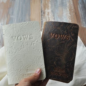 May include: Two leather-bound vow books. One is off-white, the other is dark brown. Both feature embossed floral designs and the word "VOWS". The brown book is personalized with a name and date.