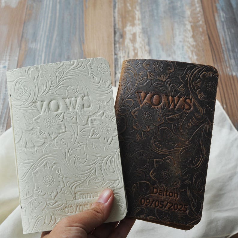 May include: Two leather-bound vow books. One is off-white, the other is dark brown. Both feature embossed floral designs and the word "VOWS". The brown book is personalized with a name and date.