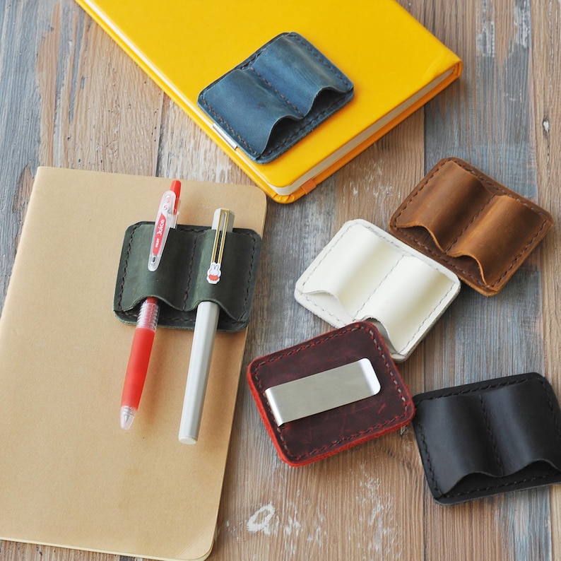 May include: Assortment of leather pen holders in various colors, including brown, black, white, and green. Some holders have two slots for pens, while others have a single slot and a money clip.