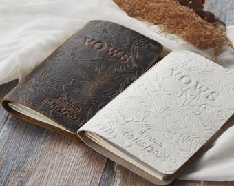 May include: Two leather-bound vow books with floral embossed designs. The brown book has the text "Vows" and "Dalton 05/05/2025" embossed on the cover. The white book has the text "Vows" and "Emma 05/05/2025" embossed on the cover.