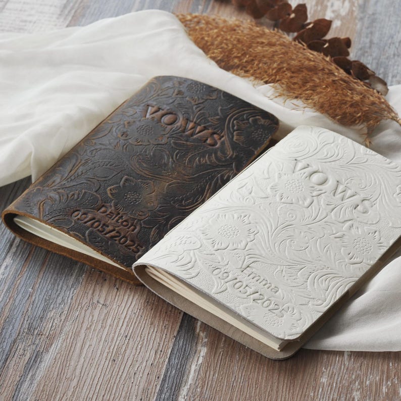 May include: Two leather-bound vow books, one brown and one white, with floral embossing. The word "VOWS" is embossed on the cover of each book. The brown book has the name "Dalton" and the date "09/05/2025" embossed on the cover. The white book has the name "Emma" and the date "09/05/2025" embossed on the cover.