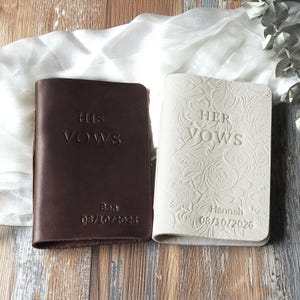 May include: Two leather vow books, one dark brown and one cream. The brown book reads "HIS VOWS" with "Ben 08/10/2026" below. The cream book reads "HER VOWS" with "Hannah 08/10/2026" below. Both books are closed and have a simple, elegant design.