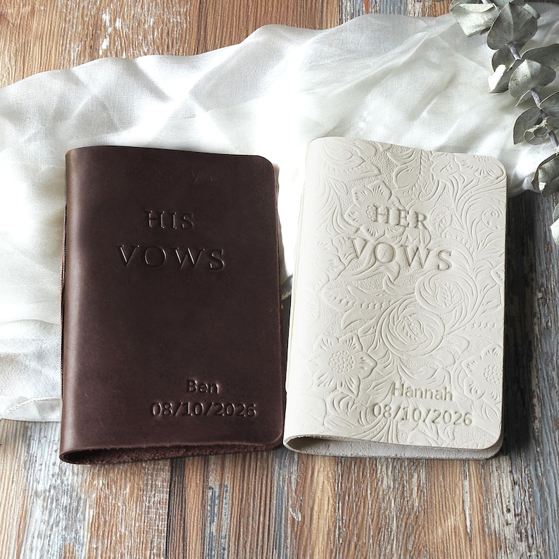 May include: Two leather vow books, one dark brown and one cream. The brown book reads "HIS VOWS" with "Ben 08/10/2026" below. The cream book reads "HER VOWS" with "Hannah 08/10/2026" below. Both books are closed and have a simple, elegant design.