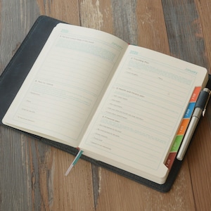 May include: Open planner with lined pages, featuring sections for planning, including a travel plan, health and fitness plan, and financial plan. The planner has colorful tabs and a pen. The cover is black, and the pages are cream-colored.
