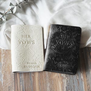 May include: Two wedding vow books, one cream and one black, with floral embossed designs. The cream book reads "HER VOWS" and the black book reads "HIS VOWS". Both books have names and dates embossed on them.