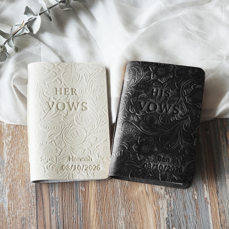 May include: Two wedding vow books, one cream and one black, with floral embossed designs. The cream book reads "HER VOWS" and the black book reads "HIS VOWS". Both books have names and dates embossed on them.