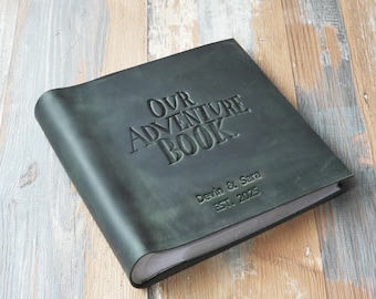 Peut inclure: Un livre relié en cuir vert foncé intitulé "OUR ADVENTURE BOOK" avec des lettres en relief. Le livre est personnalisé avec les noms "Devin & Sara" et l'année "EST. 2025". Le livre est posé sur une surface en bois.