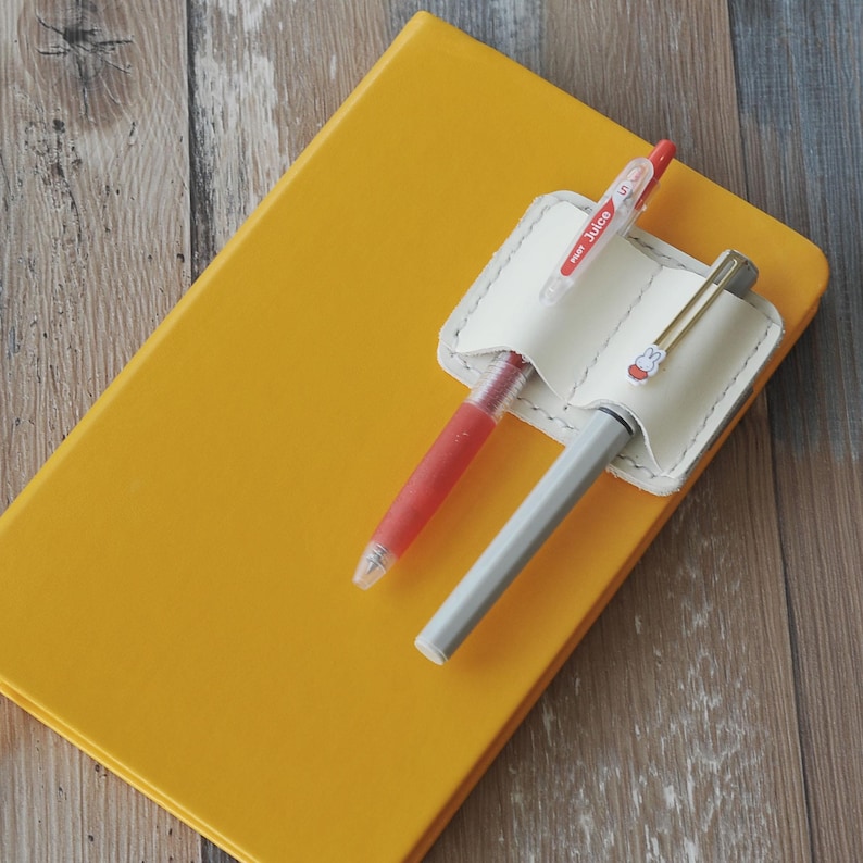 May include: A white leather pen holder with two slots for pens. The holder is attached to a yellow notebook. A red and white pen and a silver pen are in the slots.