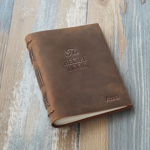 May include: A brown leather recipe book with the embossed text "The Recipe Book" and the name "Anna". The book has stitched binding and is lying on a wooden surface.