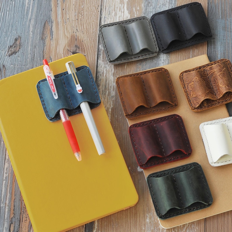 May include: A set of eight leather pen holders in various colors, including brown, red, blue, and gray. The holders are designed to hold two pens or pencils and have a flap that closes over the top. The holders are arranged in a row on a wooden surface.