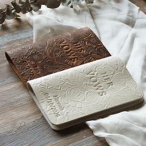 May include: Two leather vow books, one brown and one cream, with floral embossing. The brown book has the word "VOWS" and the date "08/10/2020" embossed. The cream book has the words "OUR VOWS" and the date "08/10/2026" embossed.