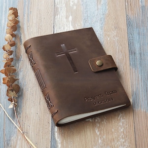 Könnte beinhalten: Ein braunes Lederjournal mit einem geprägten Kreuz auf dem Einband. Das Journal hat einen Riemenverschluss mit einem Metallknopf. Der Text "Prayers from JACKSON" ist auf dem Einband geprägt. Das Journal liegt auf einer Holzoberfläche.