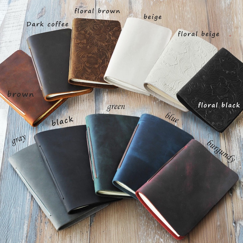 May include: A collection of leather journals in various colors, including brown, black, gray, green, and blue. Some journals feature embossed floral patterns. The journals are open, revealing the pages.