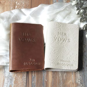 May include: Two leather vow books, one brown and one cream. The brown book reads "HIS VOWS" with the name "Ben" and the date "08/10/2026". The cream book reads "HER VOWS" with the name "Hannah" and the date "08/10/2026".