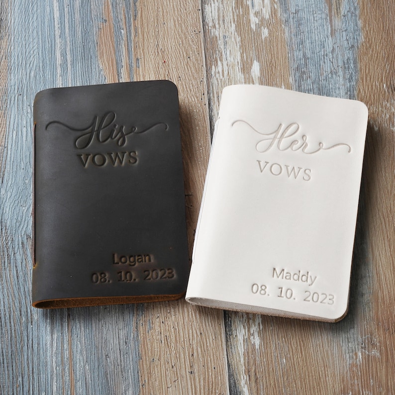 May include: Two leather-bound vow books, one black and one white. The black book has "His Vows" embossed on the front, with "Logan 08.10.2023" below. The white book has "Her Vows" embossed on the front, with "Maddy 08.10.2023" below.