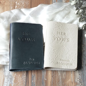 May include: Two leather vow books, one navy blue and one cream. The navy book reads "HIS VOWS" with the name "Ben" and the date "08/10/2026". The cream book reads "HER VOWS" with the name "Hannah" and the date "08/10/2026".